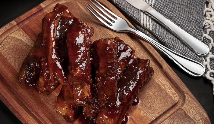 Foto de cima de uma tábua de madeira com a receita de costelinha suína ao molho barbecue, com alguns talheres de metal, um pote com mais molho e um tecido em tom cinza claro