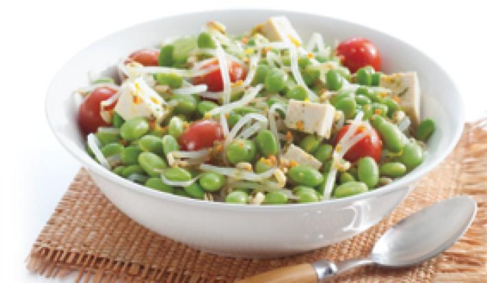 Fotografia em tons de marrom em um fundo branco com um paninho marrom e um prato fundo redondo branco com a salada de edamame, com soja verde, tomate e tofu.