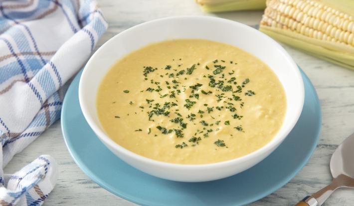 Fotografia em tons de amarelo e azul em uma bancada de madeira clara, um prato azul e branco ao centro com sopa de milho. Ao lado uma espiga de milho e um paninho azul quadriculado.