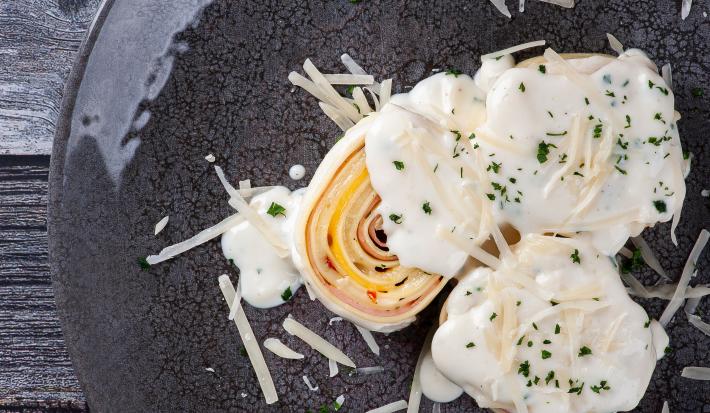 Fotografia vista de cima de três unidades de rondelli deitados em um prato raso escuro. Por cima das massas tem molho branco com queijo parmesão ralado. O prato está sobre uma mesa de madeira preta.