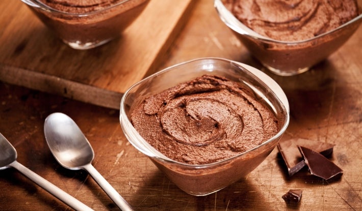 Fotografia em tons de marrom com três potes de vidro ao centro. Em cima de cada pote existe uma receita de mousse de chocolate com chia e ninho adulto.