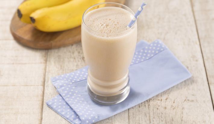 Foto em tons de bege com o copo de vitamina de banana com caramelo em cima de um paninho azul com bordas de bolinhas brancas, esses em cima de uma mesa de madeira. Fundo decorado com duas bananas nanicas em cima de uma tábua de madeira em formato oval.