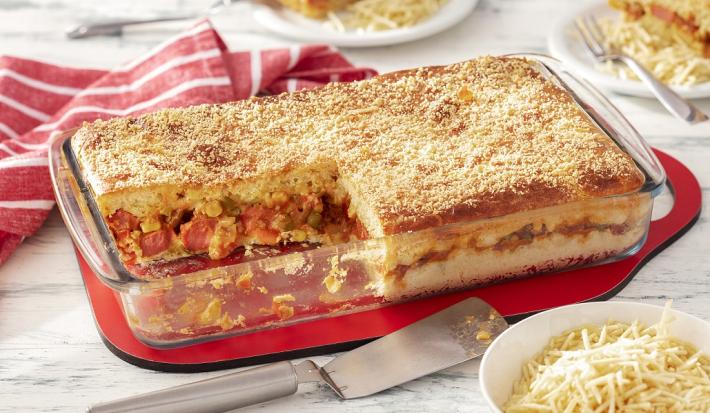 Fotografia em tons de vermelho em uma bancada de madeira de cor branca. AO centro, um pano vermelho contendo uma travessa de vidro com o cachorro quente. Ao lado há um pano listrado vermelho, um recipiente com batata palha.