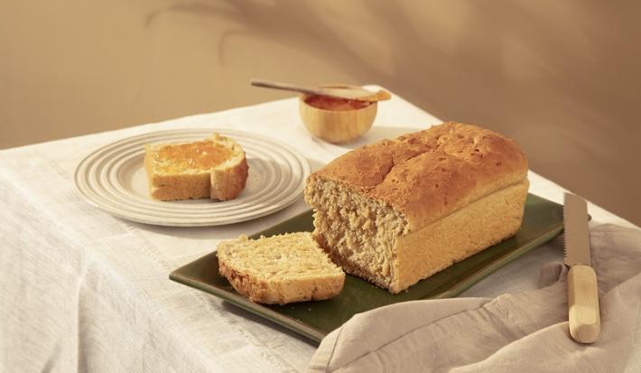 Foto em tons de bege da receita de pão com farinha de aveia servida sobre uma base de porcelana verde-escuro cortada a uma fatia com uma faca de cozinha ao lado. Ao fundo há outra fatia do pão servida em um prato bege claro e um pote de madeira com mel