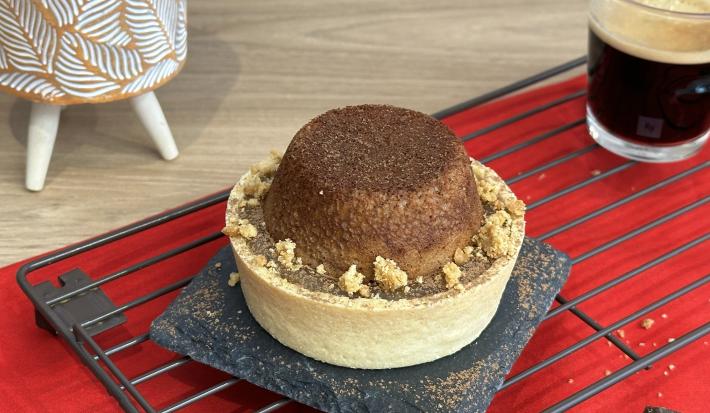 Fotografia de uma mini tortinha com base de biscoito maisena com um pequeno bolo de chocolate ao topo. Ao fundo existe um pequeno copo com café e embaixo uma tolha de cor vermelho escuro