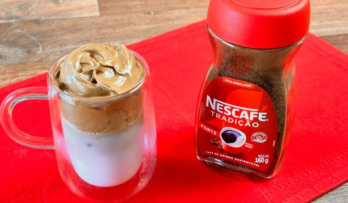 Foto aproximada da receita de Dalgona Café feito com Nescafé Tradição, servido em um xícara grande transparente ao lado de um pote de Nescafé, tudo sobre um pano vermelho em uma tábua de madeira