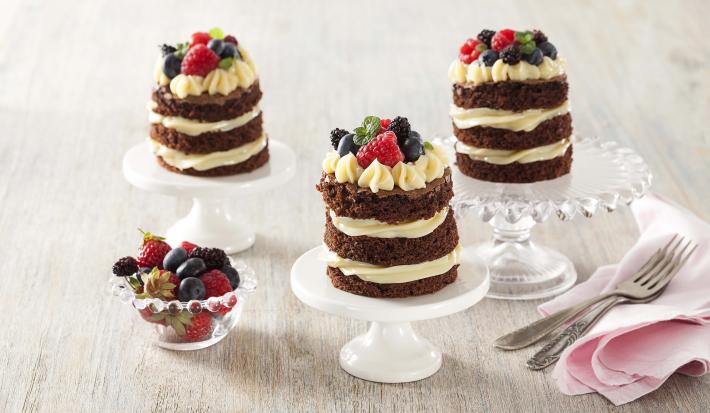 Fotografia em tons de branco e cinza de uma bancada cinza com com suportes de bolos brancos e de vidro com naked cakes. Ao lado um paninho rosa com garfos e um recipiente redondo de vidro com frutas vermelhas.