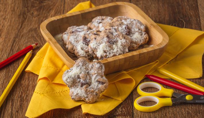 Fotografia em tons de amarelo em uma bancada de madeira de cor marrom. Ao centro, um pano amarelo contendo uma travessa de madeira em cima com os biscoitinhos dentro. Ao redor, há uma tesoura amarela e alguns lápis de cor espalhados.