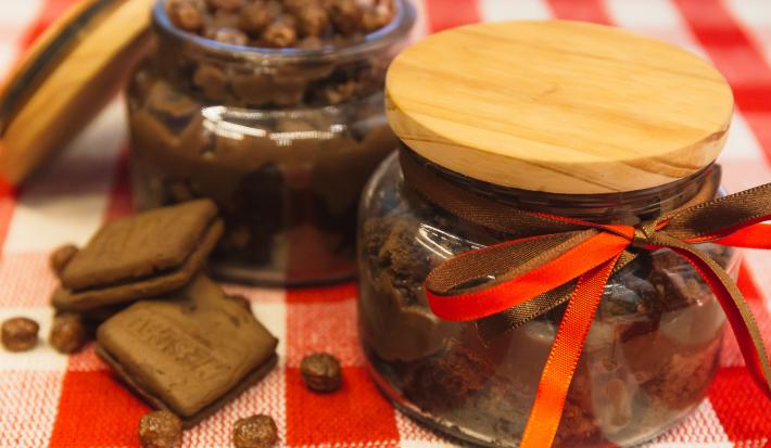 Foto de uma bancada forrada com uma toalha quadriculada vermelha e branca. Sobre ela há 2 potes de vidros com a receita de Brownie de Nescau com Brigadeiro. Um deles está fechado e o outro aberto. Além disso há 2 biscoitos e algumas bolinhas de cereal