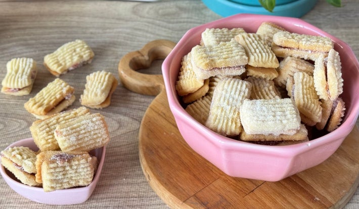 Fotografia mostra um bowl rosa com biscoitinhos retangulares. O bowl está posicionado sobre uma tábua de madeira.