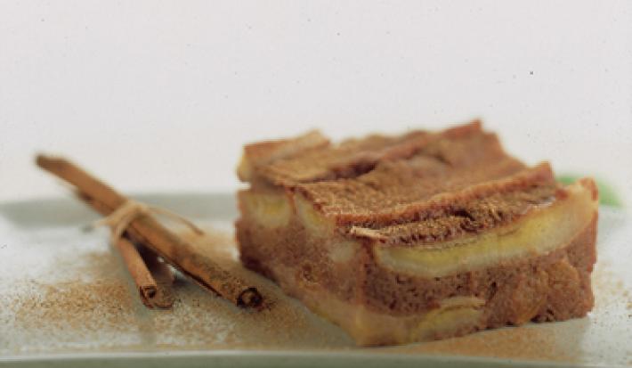 Fotografia em tons de canela e cinza, com pedaço de torta de banana com canela sobre travessa verde com ramos de canela ao lado, tudo polvilhado com canela, em fundo cinza.