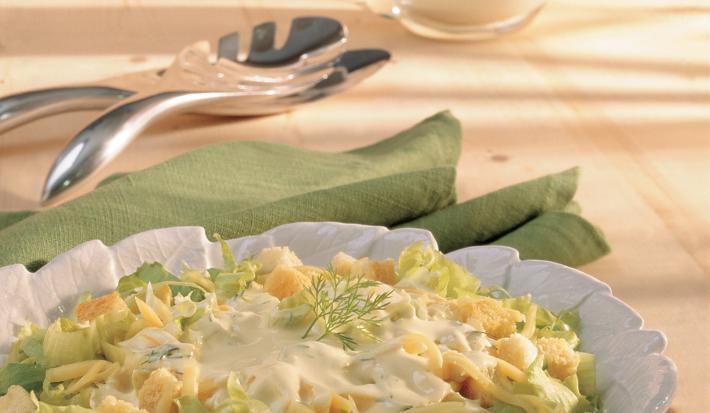 Fotografia em tons de verde em uma bancada de madeira clara, um pano verde, um pegador de salada e um prato redondo branco fundo com a salada de alface, queijo, torradinhas e iogurte dentro. Fotografia em tons de verde em uma bancada de madeira clara, um pano verde, um pegador de salada e um prato redondo branco fundo com a salada de alface, queijo, torradinhas e iogurte dentro.