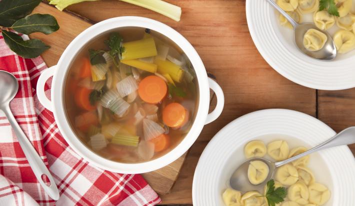 Imagem vista de cima de uma panela com caldo de legumes com pedaços de cebola, cenoura. alho poró e ao lado dois pratos de massas.