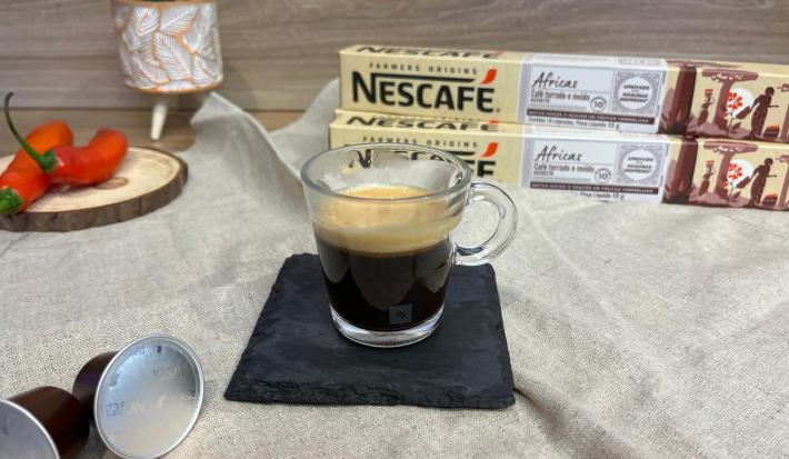Fotografia em tons de vermelho com uma xícara de vidro ao centro. Dentro da xícara existe uma dose de café espresso. Ao fundo existe duas pimentas inteiras e duas caixas de cápsulas NESCAFE