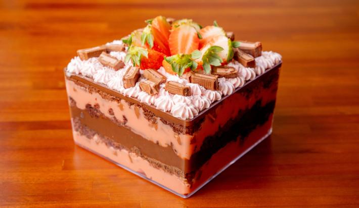 Fotografia de uma sobremesa dentro de uma travessa quadrada e transparente. Observa-se camadas de chocolate intercaladas com camadas de um creme de cor rosa. Ao topo existe uma decoração com base de chantilly, pedaços de chocolate e morangos