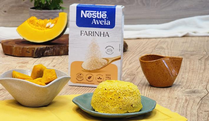 Fotografia em tons de amarelo com um prato azulado ao centro. Em cima do prato existe um pão de cor amarelada feito com abóbora e farinha de aveia. Ao fundo existe uma fatia de abóbora e uma embalagem de Farinha de Aveia Nestlé