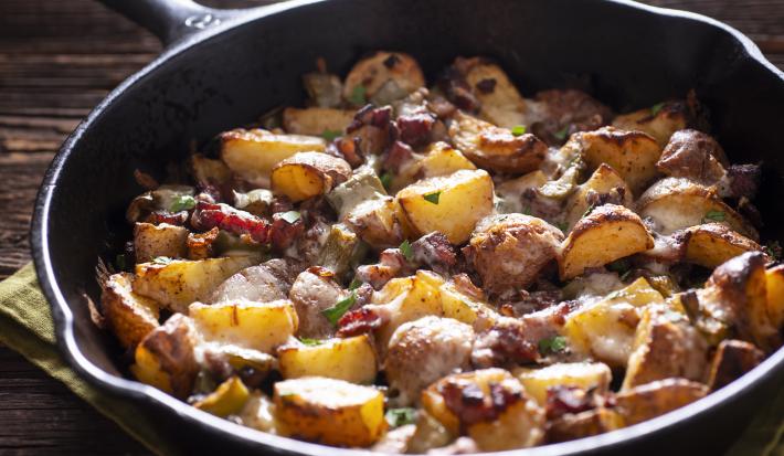 Fotografia de batatas assadas em pedaços com carne em uma panela de ferro, com salsinha por cima. A panela está apoiada em um pano verde, que está sobre uma mesa de madeira.