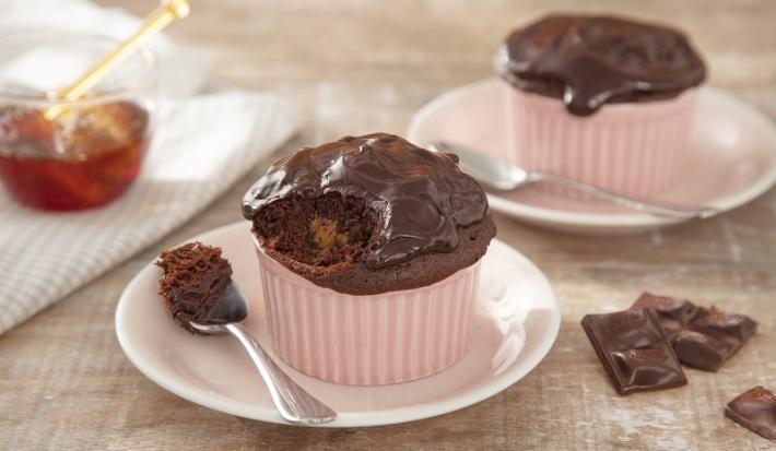 Foto em tons de marrom e cor de rosa da receita de pão de mel de micro-ondas servida em duas porções em potinhos de porcelana rosa sobre um pires branco com uma colher prateada. Ao lado há um vidro de mel sobre um pano branco