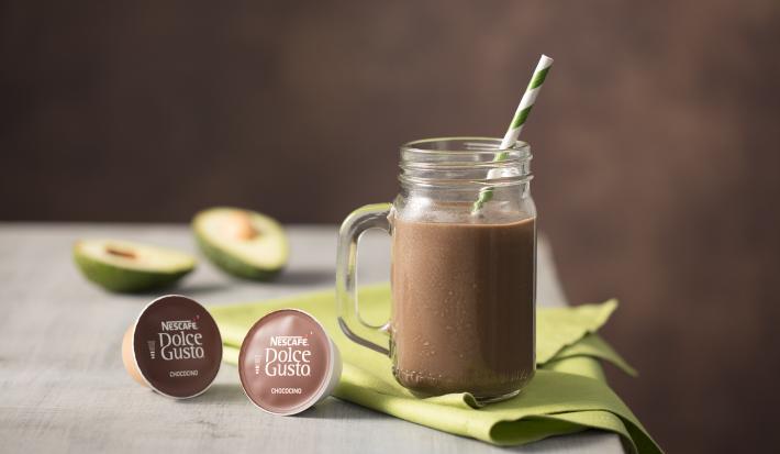 fotografia em tons de cinza e marrom de uma bancada vista de frente, contém um pano verde com uma xicara transparente com a bebida de Choccoccino Cremoso, duas capsula de Dolce Gusto, e ao fundo duas fatias de abacate