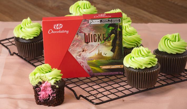 Imagem de cupcakes de chocolate com cobertura verde e um cupcake ao meio com recheio rosa. Ao lado, uma caixa de  KitKat Wicked.