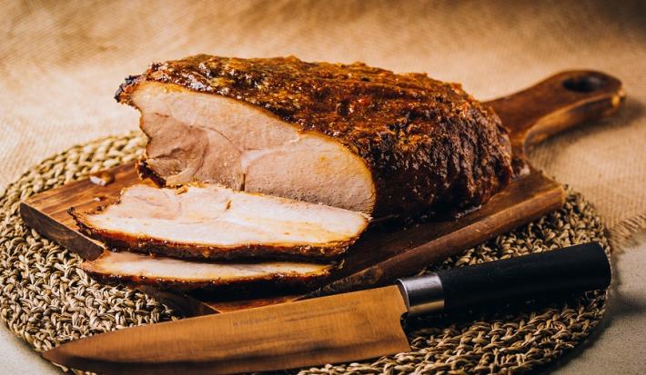 Fotografia em tons de marrom em uma bancada de madeira de cor marrom. Ao centro, uma tábua de madeira contendo o pernil e ao lado há uma faca.