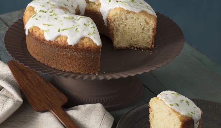 foto em tons de cinza, em uma mesa de madeira contém o bolo de iogurte ao lado um prato marrom com um pedaço de bolo um pano branco e por cima do pano uma espátula.