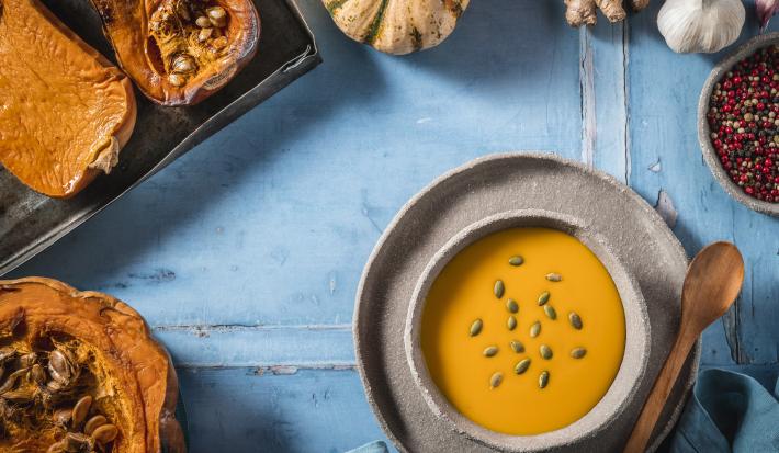 Fotografia vista de cima de um recipiente fundo de cerâmica com uma sopa de abóbora sobre um prato de cerâmica raso, com uma colher de madeira apoiada. Ao redor, várias abóboras assadas, pimentas em grãos e uma cabeça de alho, sobre uma mesa azul.