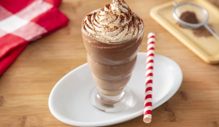 Foto da receita de Milkshake de Cappuccino. Observa-se um copo alto com a bebida decorado com chantilly e polvilhado de chocolate em pó. Ao lado direito, um canudo vermelho e branco para decorar.