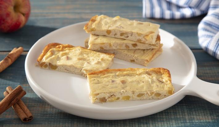 Foto em tons de amarelo da receita de barrinha proteica de cheesecake de maçã servida em diversas porções sobre uma panela branca de cerâmica em cima de uma mesa de madeira azul com paus de canela e maçã