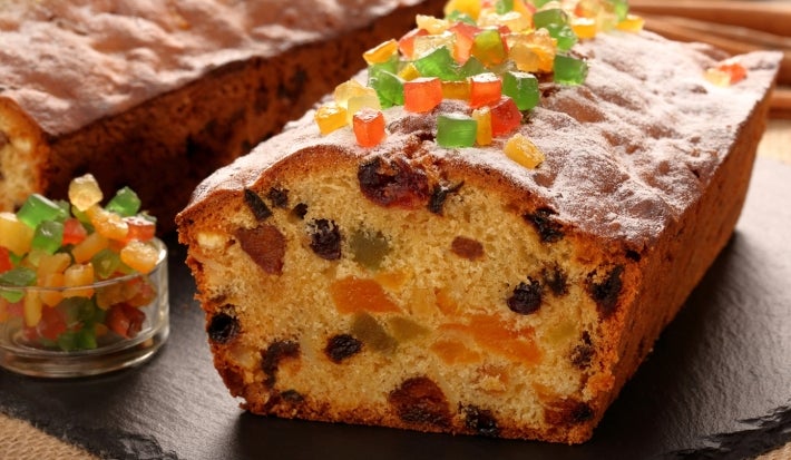Em cima de uma mesa, bolo fofinho com frutas cristalizadas e uvas-passas, deccorado com frutas cristalizadas.
