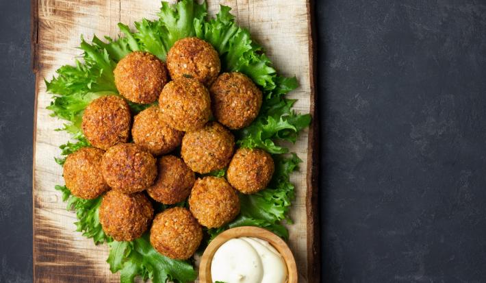 Fotografia vista de cima de várias unidades de falafel sobre uma folha de alface e um apoio de madeira retangular, ao lado um molho de iogurte que também está sobre o apoio.