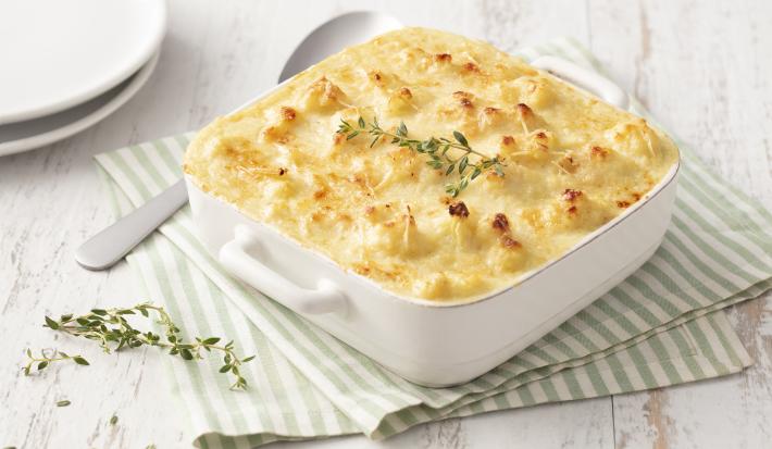 Fotografia em tons de branco e verde de uma bancada de madeira branca com um paninho listrado branco e verde, sobre ele um recipiente quadrado branco com o gratinado e uma colher. Ao fundo dois pratos brancos.