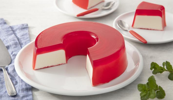 Fotografia em tons de rosa em uma bancada de madeira clara, um paninho azul, um prato branco redondo raso com a gelatina de morango recheada com creme branco. Ao fundo, pratinhos brancos com fatias da gelatina e talheres com cabo vermelho.