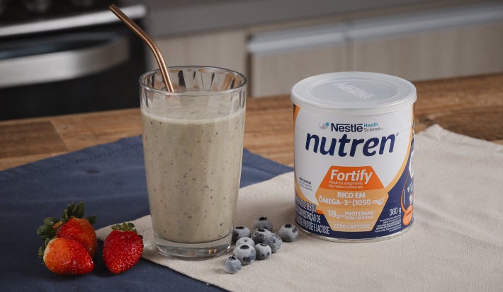 A imagem mostra a receita da vitamina fortificante servida em um copo de vidro alto e largo com um canudo dourado, ao lado há morangos, mirtilos e uma lata de nutren fortify