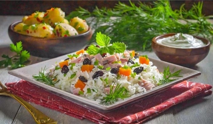 Em um prato quadrado de porcelana, há uma base de arroz soltinho, combinado com pedaços de presunto cozido, coloridos vegetais, como cenoura, uvas-passas e decorado com cebolinha picada.