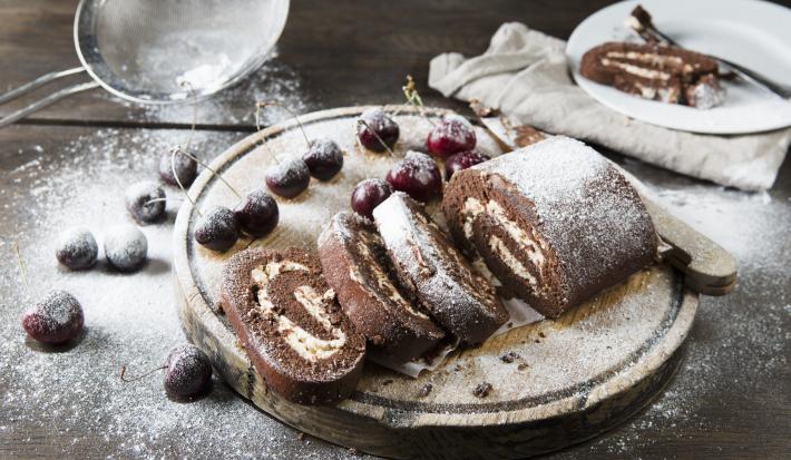 Fotografia de um rocambole de pão-de-ló de chocolate, recheado com Leite MOÇA, coco e castanha-de-caju. Por cima tem um povilhado de açúcar de confeiteiro