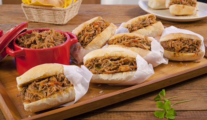 Fotografia em tons de marrom, vermelho e branco de uma bancada de madeira com uma tábua de madeira, sobre ela pães recheados com pernil desfiados dentro de saquinhos de papel brancos. Ao lado um recipiente