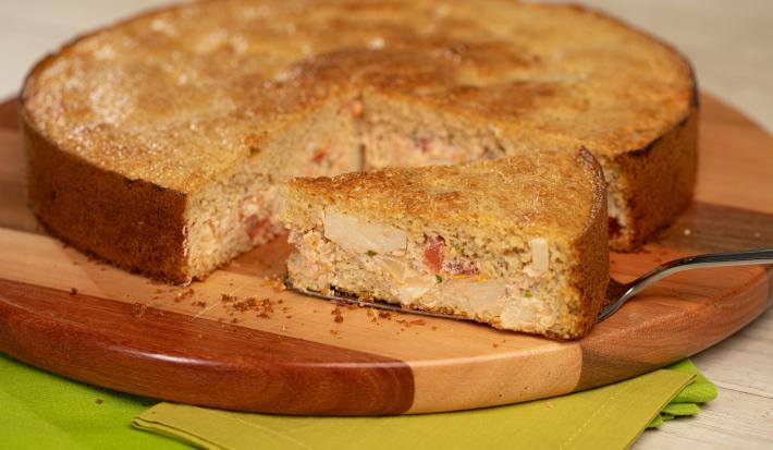 Foto de uma bancada com dois tecidos verdes, uma tábua de madeira redonda. Sobre ela, há uma torta preparada cortada com uma fatia sobre uma espátula