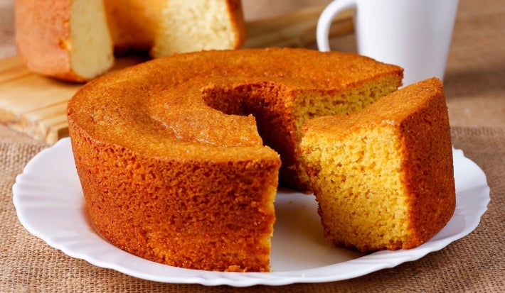 Fotografia de um bolo de milho recém-assado, com uma fatia cortada que revela seu interior macio. O bolo, em um prato com borda ondulada, com uma caneca ao fundo, transmitindo uma atmosfera caseira.