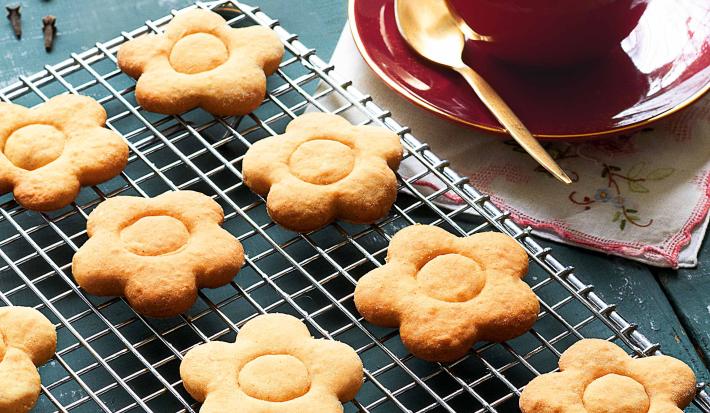 fotografia em tons de azul e bege de uma bancada azul vista de cima, contém uma grade com biscoitinhos em formato de flores e ao lado um pratinho vermelho com uma xicara vermelha que contém a bebida de chocolate branco.