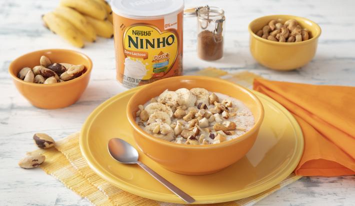 Fotografia em tons de laranja e amarelo de uma bancada de madeira, ao centro um prato amarelo e uma tigela laranja com o mingau. Ao fundo pote com castanhas, um cacho de bananas, lata de NINHO Zero Lactose e pote de canela.