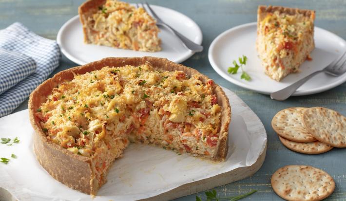 Fotografia em tons de azul e branco de uma bancada azul com um prato retangular branco, sobre ele uma torta de legumes. Ao lado biscoitos tostines. Ao fundo dois pratos com duas fatias da torta e dois garfos.