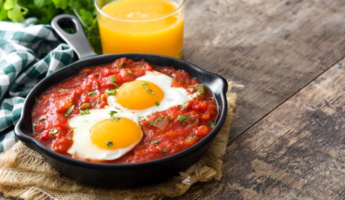 Fotografia de uma receita feita em um panela preta com molho de tomate, ovos e acompanhada de um suco de laranja