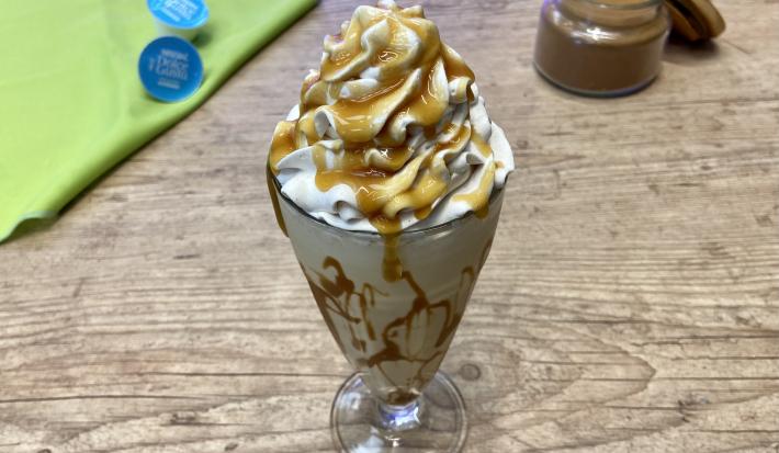 Fotografia em tons de azul e verde em uma bancada de madeira e um copo de vidro ao centro com a bebida de Milkshake e ao fundo um potinho com canela em pó.