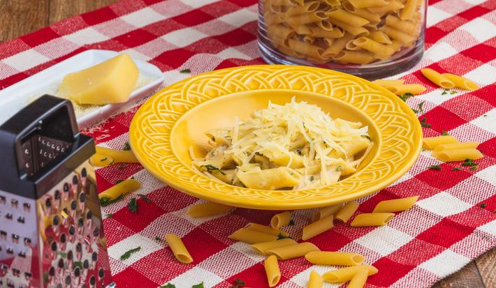 Foto de uma mesa com uma toalha xadrez vermelha e branca. Sobre ela há alguns macarrões crus, um ralador, um pedaço de queijo, um pote com mais macarrão e, ao centro, há um prato amarelo com a receita de Penne Alfredo.