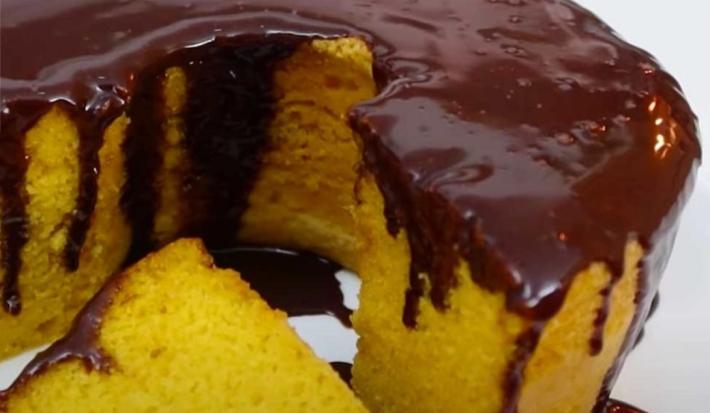 Foto da receita de Bolo de Cenoura com Cobertura de Chocolate. Observa-se um bolo com furo no meio bem de perto com uma fatia cortada e a cobertura de chocolate escorrendo