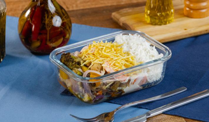 Fotografia de uma marmita retangular pequena com arroz, fricassê, batata palha, arroz e legumes assados. A marmita está sobre dois panos sobrepostos de cor azul, ao lado de talheres e uma mini tábua com pimenta e azeite.