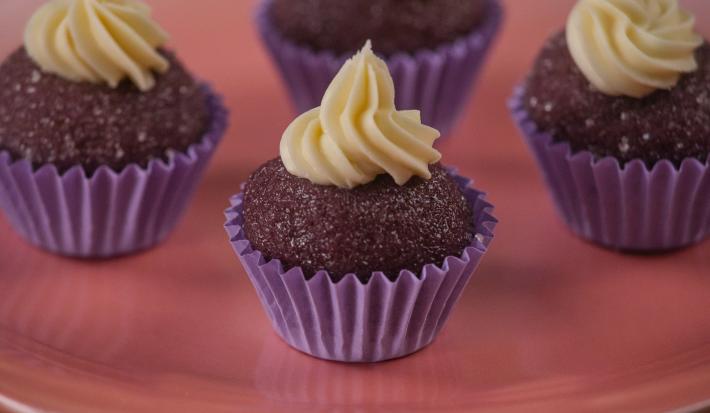 Fotografia de quatro forminhas de cor roxa com um brigadeiro de açaí, e por cima de cada um tem uma cobertura de chocolate confeitado. As unidades de brigadeiro estão sobre um apoio de cor rosa.