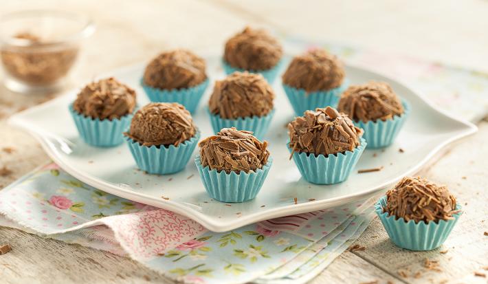 Fotografia em tons de branco, azul e marrom de uma bancada vista de frente. Panos floridos e acima um prato quadrado que contém vários brigadeiros com raspas de chocolates por cima para decorar.