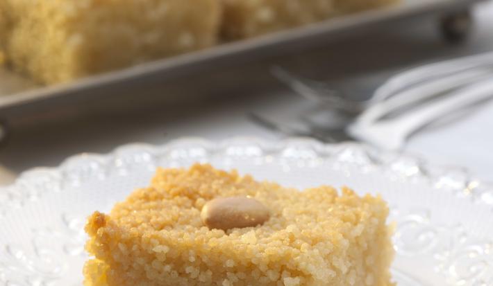 Fotografia em tons dourado e grafite, com pedaço de doce sobre prato de vidro, acima alguns garfos e bandeja grafite com outros pedaços de doce, tudo sobre toalha branca.